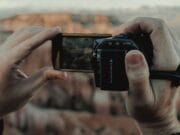 A person holds a digital camera, framing a landscape view of a canyon in the background.