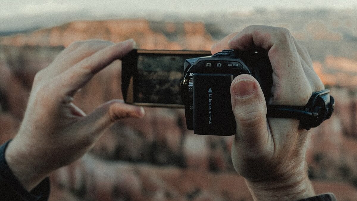 A person holds a digital camera, framing a landscape view of a canyon in the background.