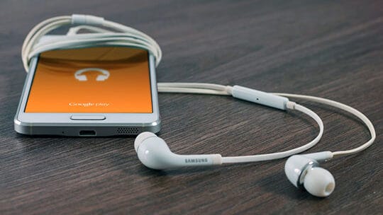 Smartphone with an orange display showing a headphone icon is placed on a wooden surface, accompanied by white Samsung earphones.