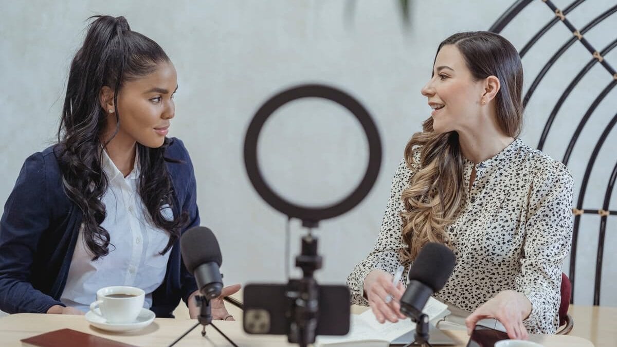 Two women sitting at a table with coffee, talking in front of microphones and a smartphone on a stand.