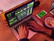 Person typing on a laptop displaying complex graphs and codes. Nearby are a smartphone, two drinks, and a bowl of chips on the table.