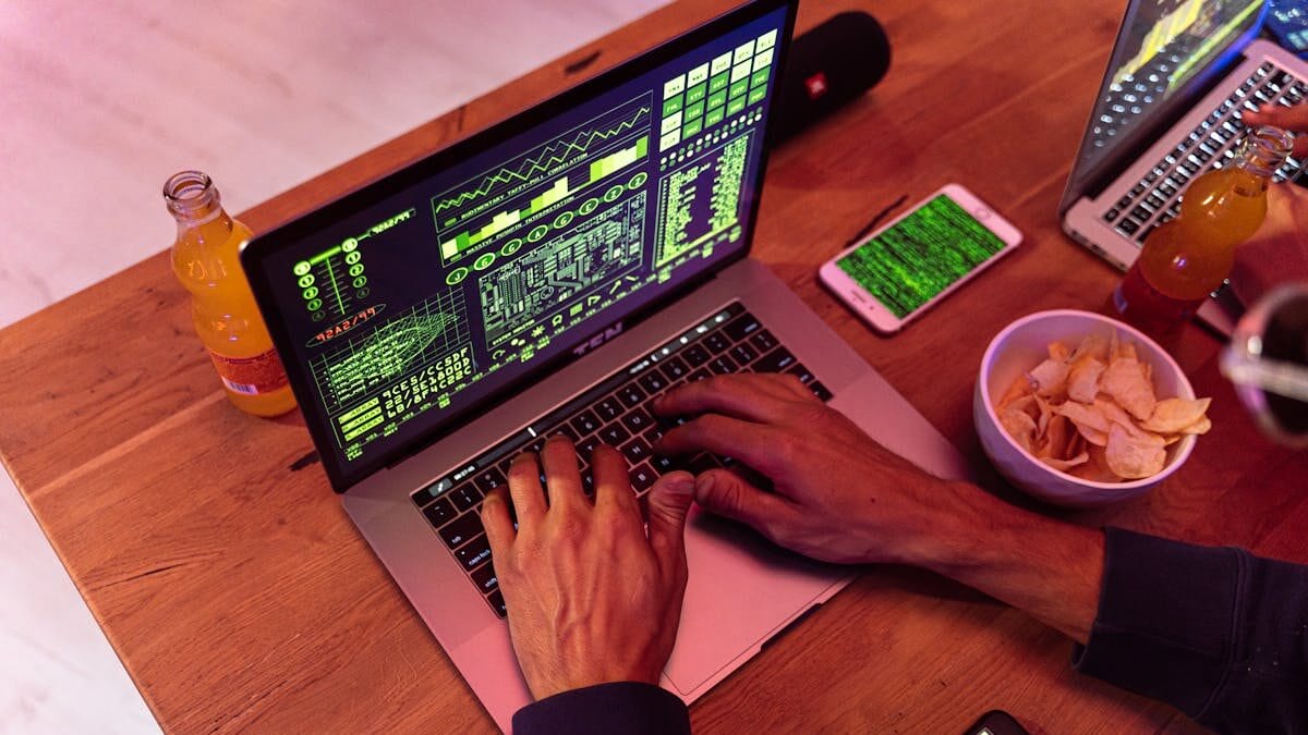 Person using a laptop with green code displayed. Nearby are a phone showing code, two bottles of juice, a bowl of chips, and another laptop, all on a wooden table.