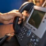 A person picking up the handset of a desk phone under a lamp.