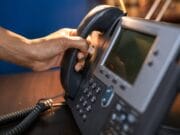 A person picking up the handset of a desk phone under a lamp.