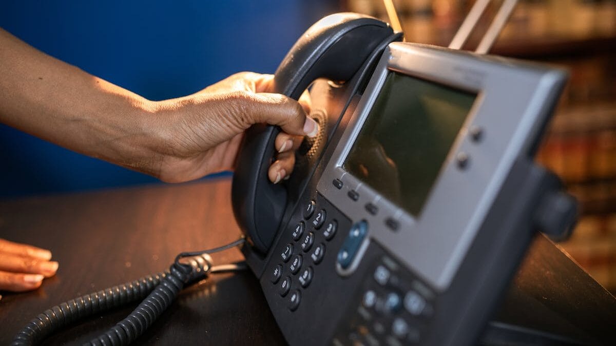 A person picking up the handset of a desk phone under a lamp.