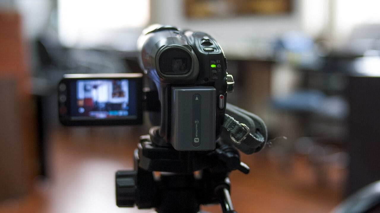 A digital camcorder on a tripod is turned on, with its display showing a scene in the background.