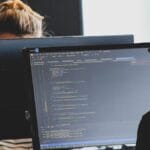 Two people work on computers in an office. The foreground screen displays code in a text editor.