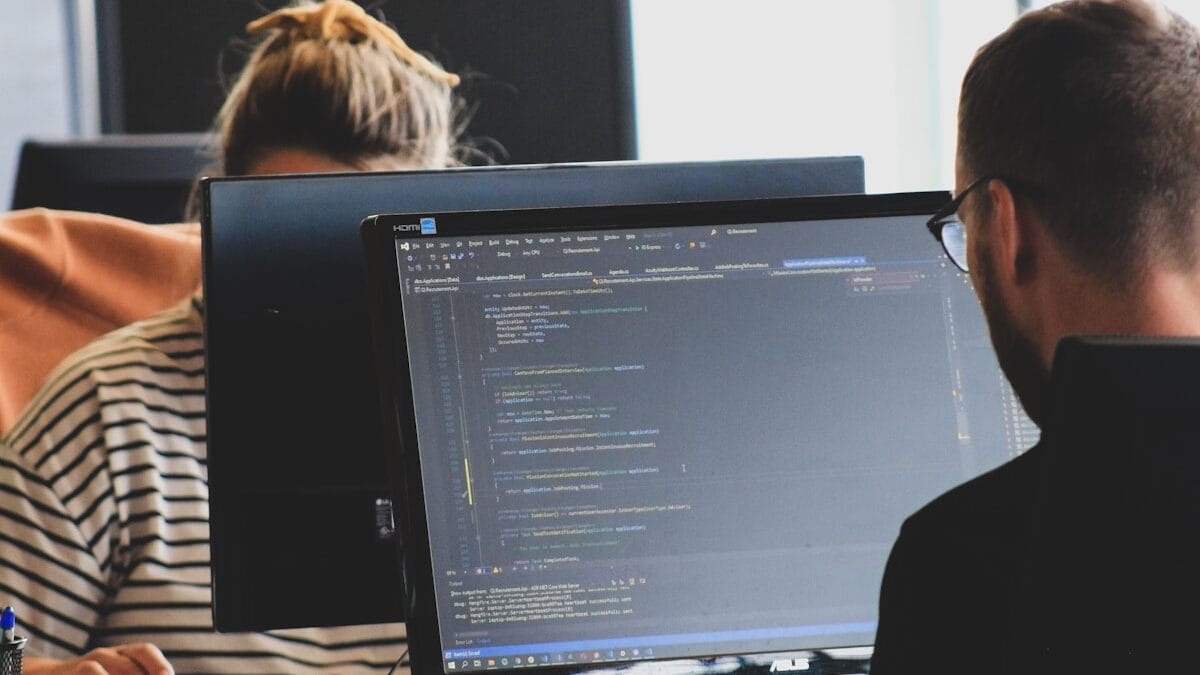 Two people work on computers in an office. The foreground screen displays code in a text editor.