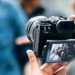 Person holding a camera, capturing an image of two people displayed on the camera's screen.