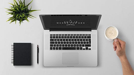 Top view of a workspace with a notebook, pen, laptop, and a hand holding a cup of coffee next to a small plant on a white surface.