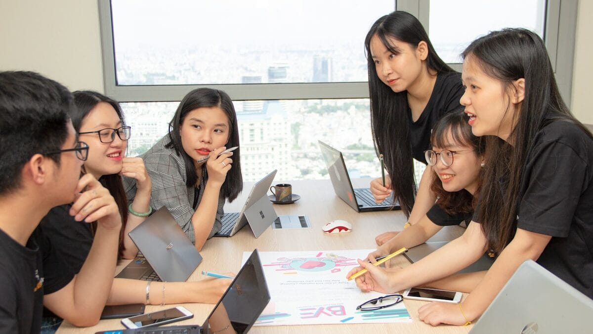 A group of six people at a table with laptops, discussing documents.