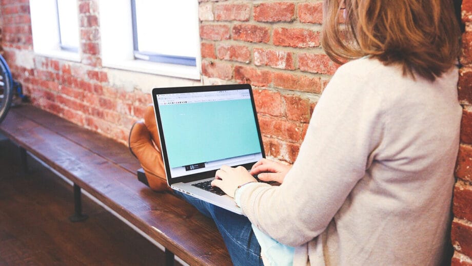 A woman sits on a wooden bench with her back against a brick wall, typing on an open laptop.