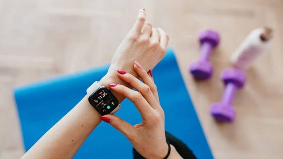 Person checks a smartwatch on their wrist while sitting on a blue exercise mat, with purple dumbbells and a bottle nearby.