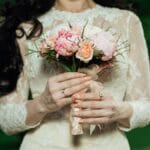 A person in a lace dress holds a bouquet of pink and peach flowers against a blurred green background.