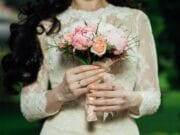 A person in a lace dress holds a bouquet of pink and peach flowers against a blurred green background.