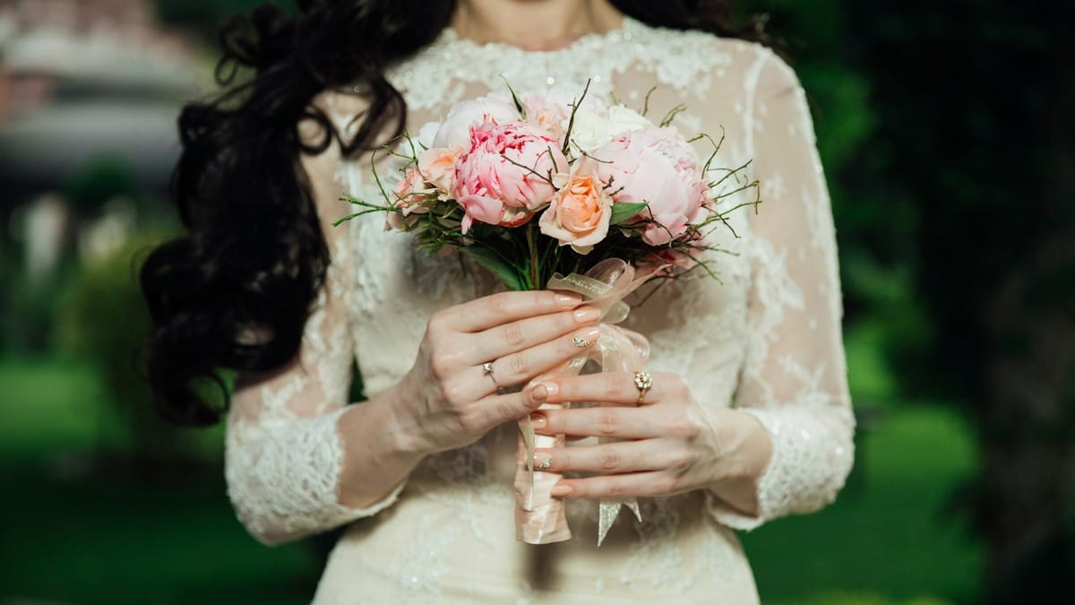 A person in a lace dress holds a bouquet of pink and peach flowers against a blurred green background.