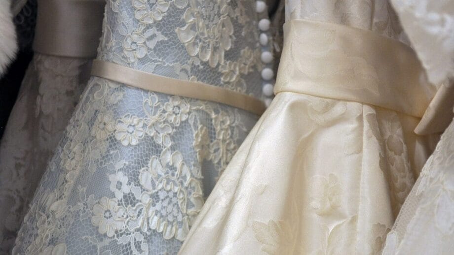 Close-up of two elegant wedding dresses on hangers, featuring intricate lace patterns and satin sashes.