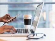 Person using a laptop and holding a smartphone. A pair of glasses and a coffee cup are on the table.