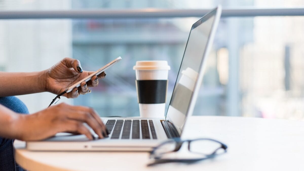 Person using a laptop and holding a smartphone. A pair of glasses and a coffee cup are on the table.