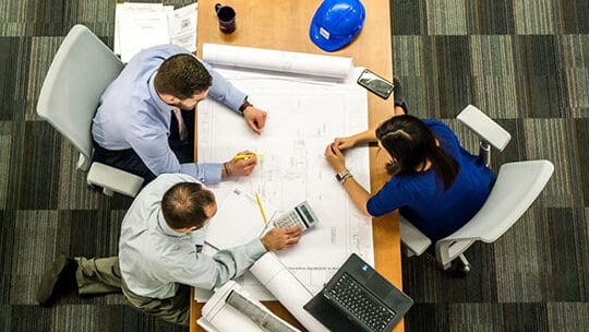 Three people reviewing architectural plans at a table with a laptop, calculator, and blue hard hat. Top-down view.
