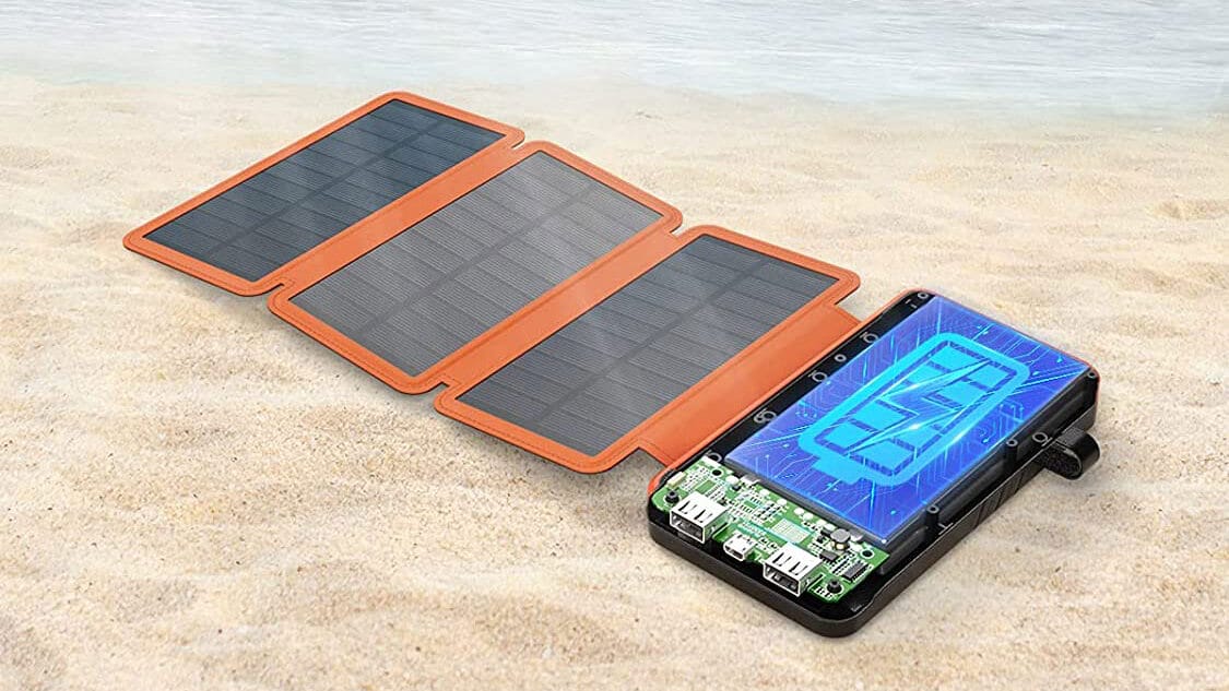 A solar charger with four foldable panels and a digital display is placed on sandy beach near the water.