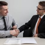 Two men sitting at a table shake hands over a document, wearing formal attire, in a professional setting.