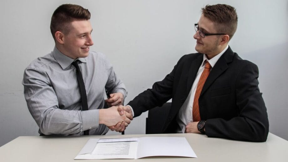 Two men sitting at a table shake hands over a document, wearing formal attire, in a professional setting.