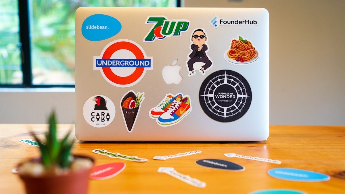 Laptop covered with various colorful stickers, including 7up, Underground, and others, on a wooden table with scattered stickers nearby; a small plant is in the foreground.