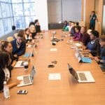 A diverse group of people sits around a long conference table with laptops and papers in a modern meeting room.