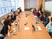 A diverse group of people sits around a long conference table with laptops and papers in a modern meeting room.