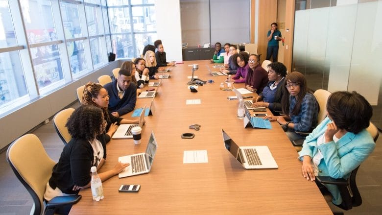 A diverse group of people sits around a long conference table with laptops and papers in a modern meeting room.