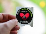 Hand holding a sticker that reads, I got my COVID-19 vaccine StopCOVID with a heart design and matrix logo.