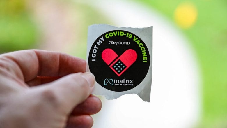 Hand holding a sticker that reads, I got my COVID-19 vaccine StopCOVID with a heart design and matrix logo.