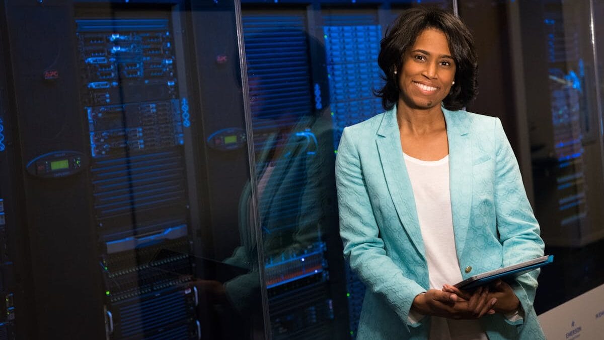 A person in a turquoise blazer stands smiling in front of server racks, holding a tablet.