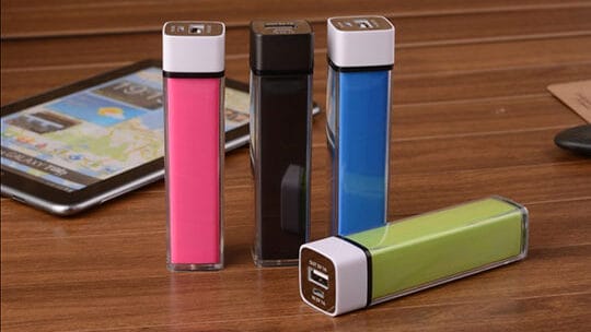 Four portable power banks in pink, black, blue, and green are standing on a wooden table, with a tablet placed nearby.