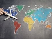 A hand holds a small toy airplane over a chalkboard world map with continents in different colors.