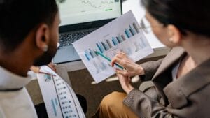 Two people reviewing printed charts and graphs over a desk with a laptop displaying financial data in the background.
