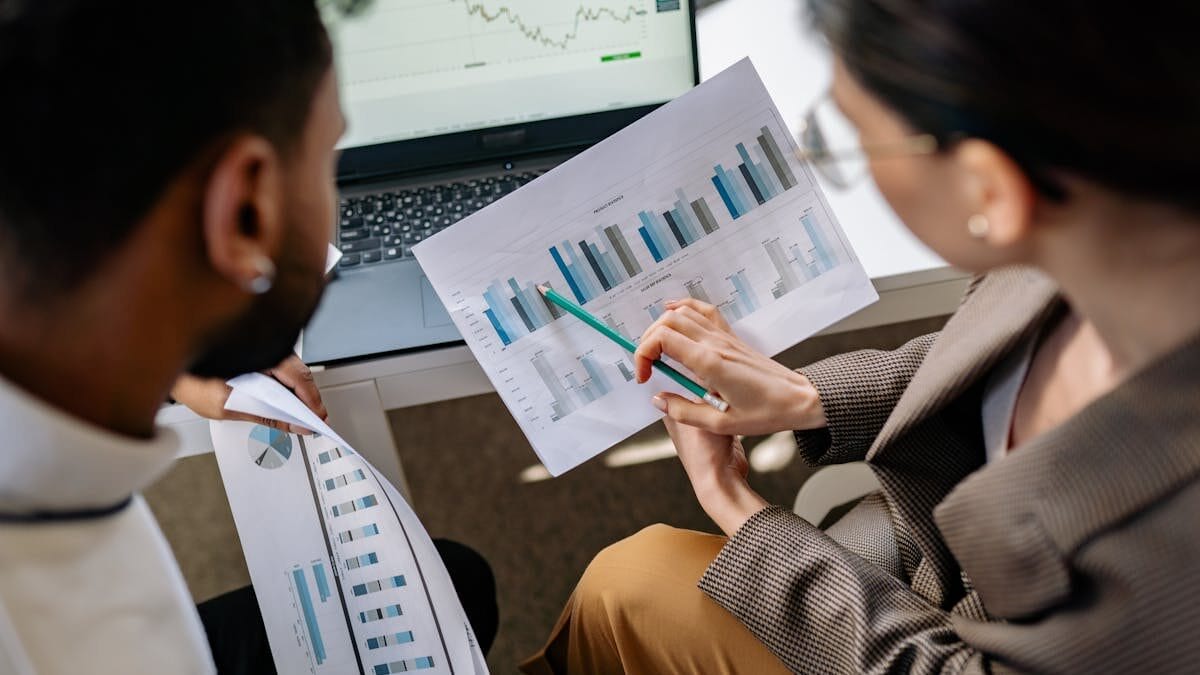 Two people reviewing printed charts and graphs over a desk with a laptop displaying financial data in the background.