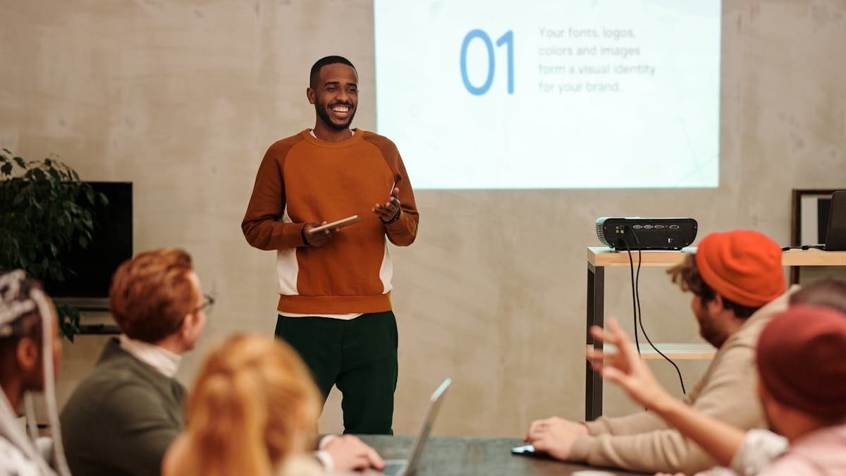 A person in a brown sweater leads a presentation in a casual meeting room, addressing seated attendees around a table.