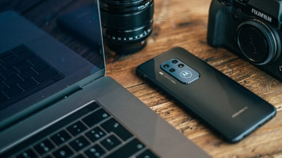 Laptop, motorola phones, fujiflim camera, and lens on a wooden table, arranged closely together.