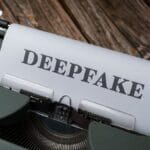 A typewriter with a sheet of paper displaying the word DEEPFAKE in bold, set on a wooden surface.