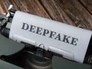 A typewriter with a sheet of paper displaying the word DEEPFAKE in bold, set on a wooden surface.