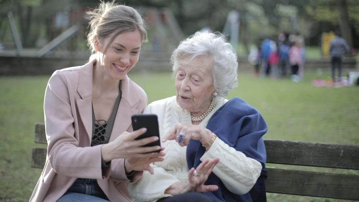 Two women sitting on a park bench, one pointing at a smartphone screen while the other smiles.