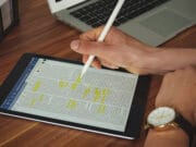 A person uses a stylus on a tablet displaying a document with highlighted text. A laptop and a wire mesh organizer are on the wooden table.