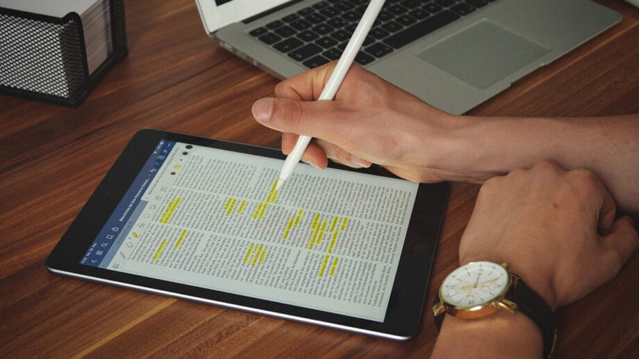 A person uses a stylus on a tablet displaying a document with highlighted text. A laptop and a wire mesh organizer are on the wooden table.