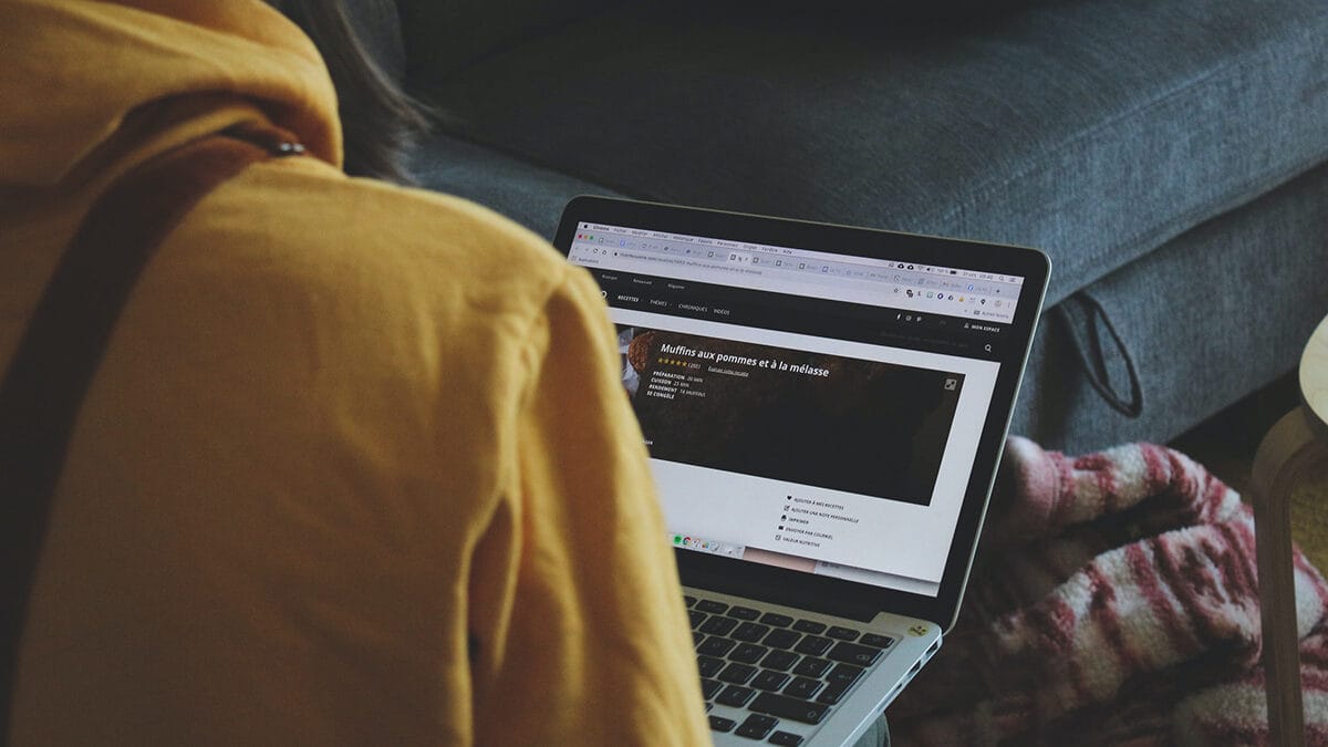 Person in a yellow hoodie using a laptop on a couch with a colorful pillow, screen showing a website.