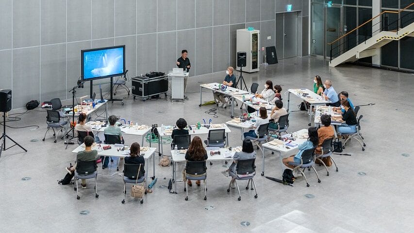 A group of people sit at tables arranged in a U-shape in a modern conference room, facing a screen with a presenter standing nearby.