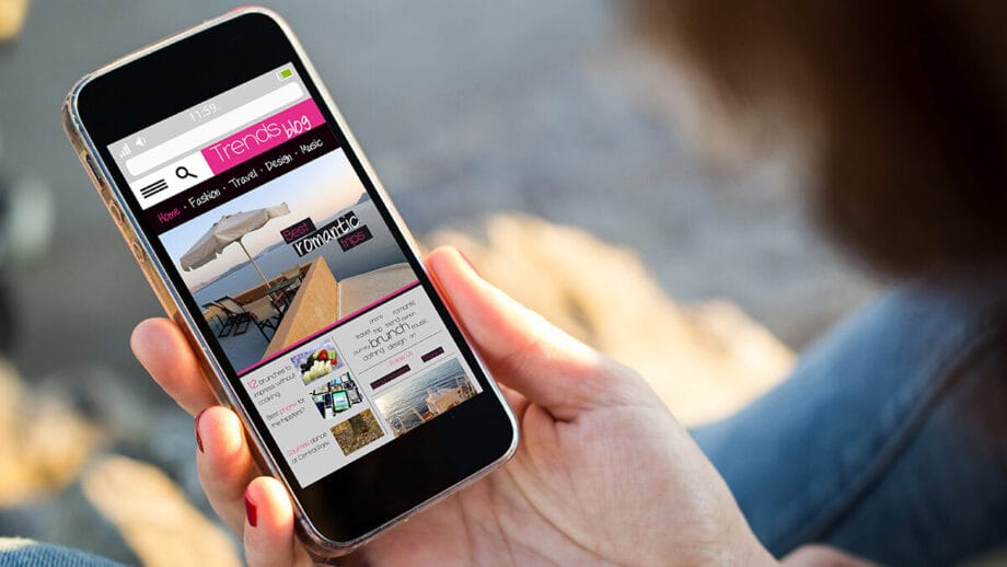 Person holding a smartphone displaying a website with categories like fashion, travel, and lifestyle. The screen shows images and text blocks.