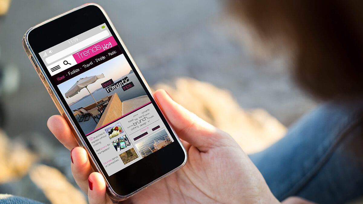 Person holding a smartphone displaying a website with categories like fashion, travel, and lifestyle. The screen shows images and text blocks.