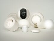 A variety of white smart home devices including a camera, speaker, plug, light bulb, and sensor are displayed against a white background.